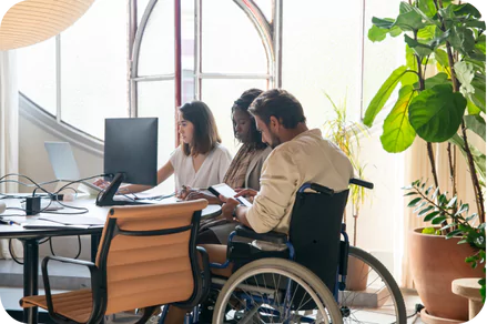 a group of three people working together at a desk in an office setting. Two women are seated at the desk, one using a laptop and the other looking at a computer monitor. A man in a wheelchair is also at the desk, holding a tablet. The office has large, arched windows that let in natural light, and there are several large potted plants around the room, creating a bright and pleasant atmosphere.