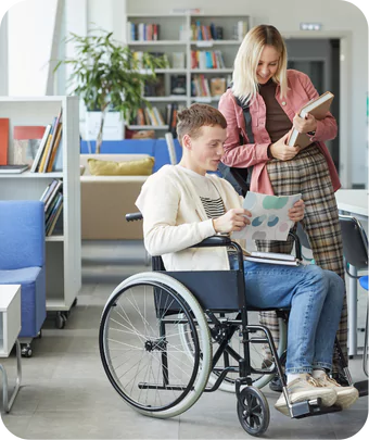 a young man sitting in a wheelchair and a young woman standing next to him in a library or study area. The man is wearing a white sweater and blue jeans, and he is holding a piece of paper with colorful circles on it. The woman is wearing a pink jacket over a beige top and plaid pants, and she is holding a book and a notebook. They both appear to be engaged in a conversation or looking at the paper together.